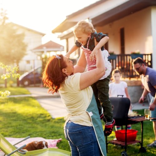 Woman is lifting up high her gorgeous and cheerful toddler boy while rest of the family is grilling.